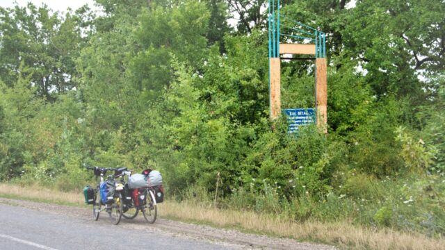 The sign behind the bushes says, "Welcome to the territorial community of Bronnytsia; settlements: Bronnytsia, Hryhorivka, Olenivka, Kryshtofivka, and Nova Hryhorivka; population: 2304; farms: 791" (Olenivka)